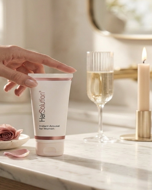 Hand holding a tube of 'HerSolution' Gel on a marble surface with a glass of champagne and candle in the background.
